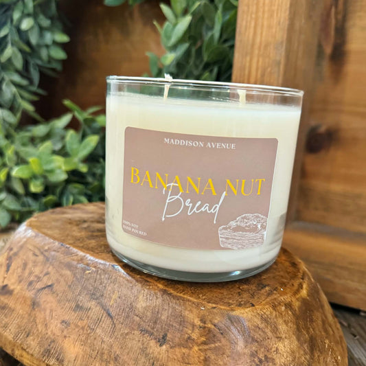 Candle labeled 'Banana Nut Bread' on a wooden surface with greenery in the background