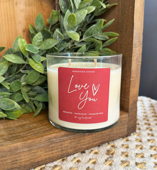 Candle with a 'Love You' label in front of greenery on a wooden surface
